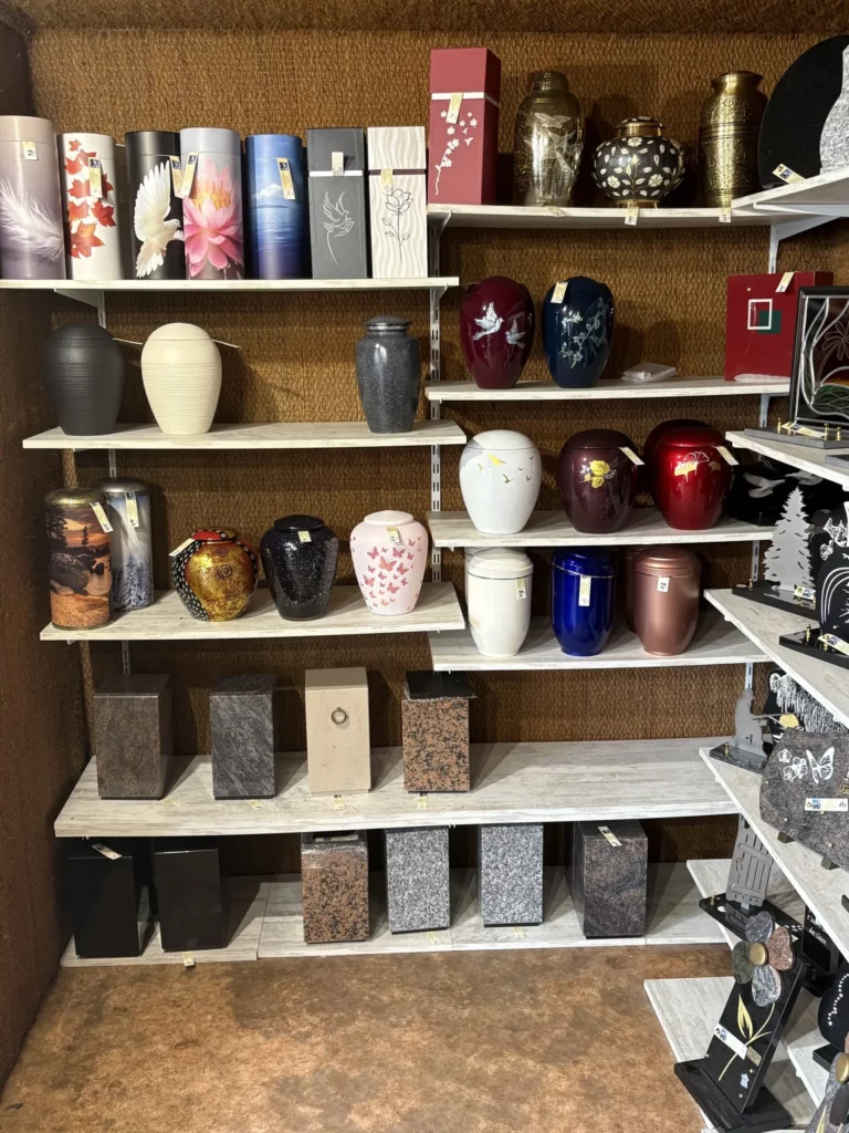 Collection d'urnes cinéraires aux styles et matériaux variés sur des étagères blanches.