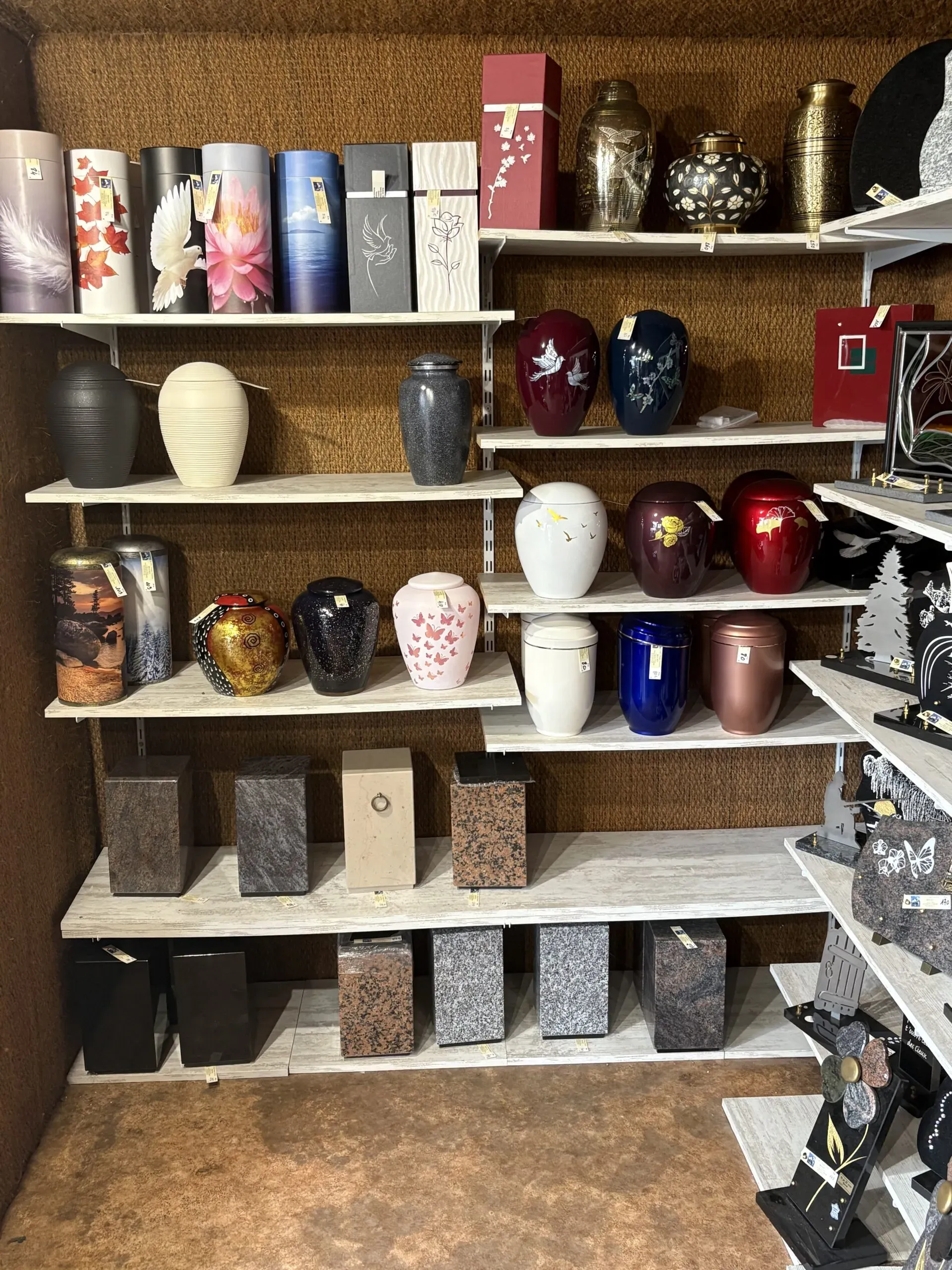 Collection d'urnes cinéraires aux styles et matériaux variés sur des étagères blanches.