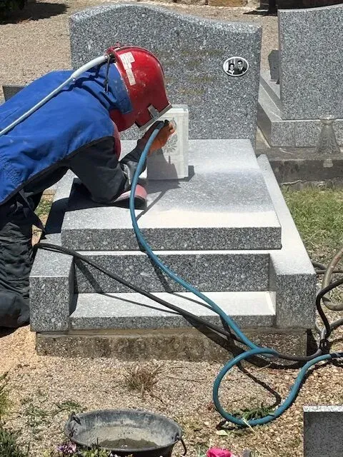 Marbrier en casque rouge et combinaison bleue effectuant un sablage au cimetière.